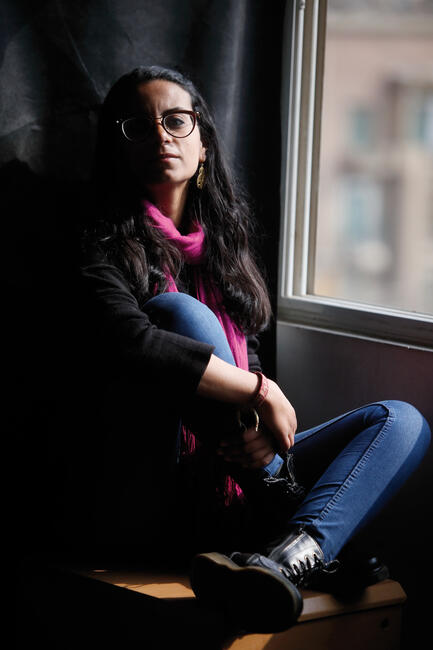 Eine junge Frau mit Brille, Jeans und schwarzen Stiefeln sitzt neben einem Fenster