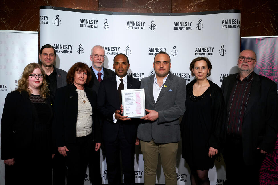 Menschen stehen vor einer Pressewand mit Amnesty-Logo, zwei von ihnen halten einen Preis