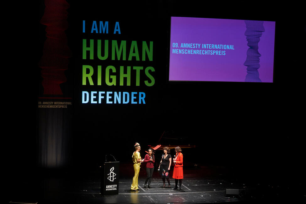 Vier Frauen sprechen auf einer großen Bühne vor der Hintergrundprojektion "I am a human rights defender"