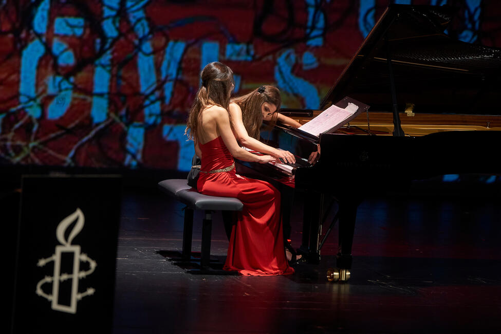 Zwei Frauen in langen Abendkleidern spielen gemeinsam an einem schwarzen Klavierflügel auf einer Bühne