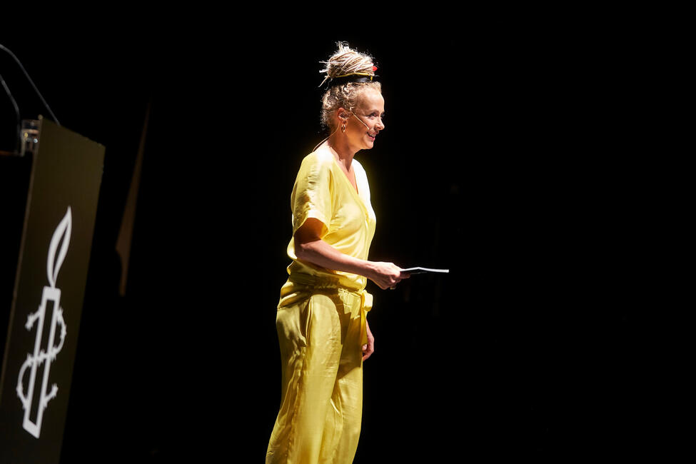 Eine Frau in gelber Abendkleidung und Headset-Mikro steht auf einer Bühne