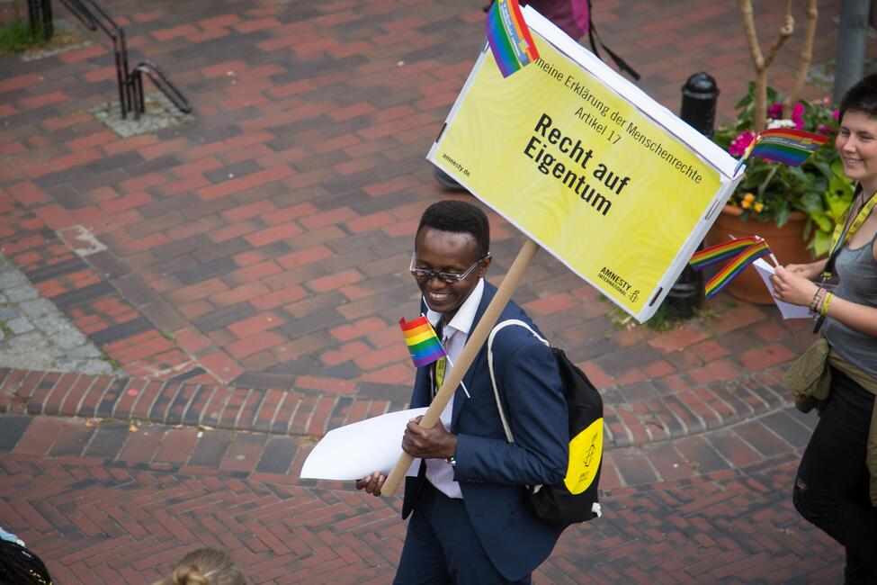 Ein schwarzer Mann mit Sonnenbrille und im blauen Sakko trägt ein gelbes Schild auf dem steht: "Recht auf Eigentum"