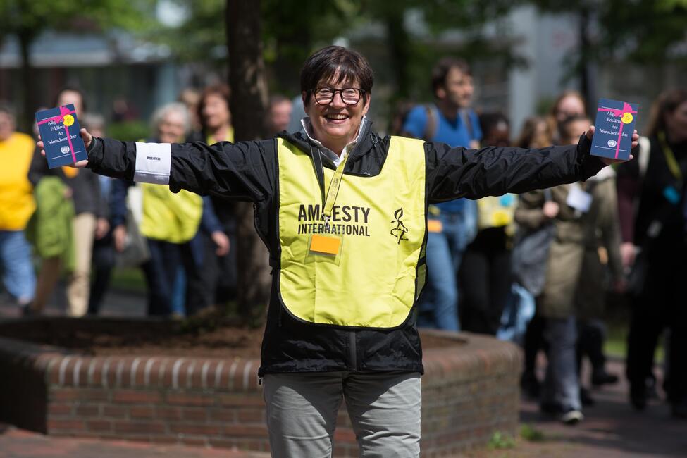 Eine Frau in einer gelben Weste, auf der Amnesty International darauf steht, hält in beiden Händen die Büchlein der "Allgemeinen Erklärung der Menschenrechte"