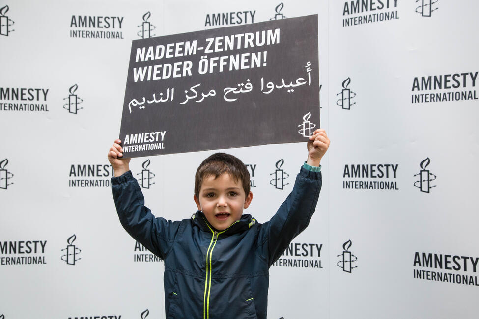 Ein kleiner Junge steht vor einer Amnesty-Fotowand und hält ein Schild in die Luft, auf dem steht:"Nadeem-Zentrum wieder öffnen"