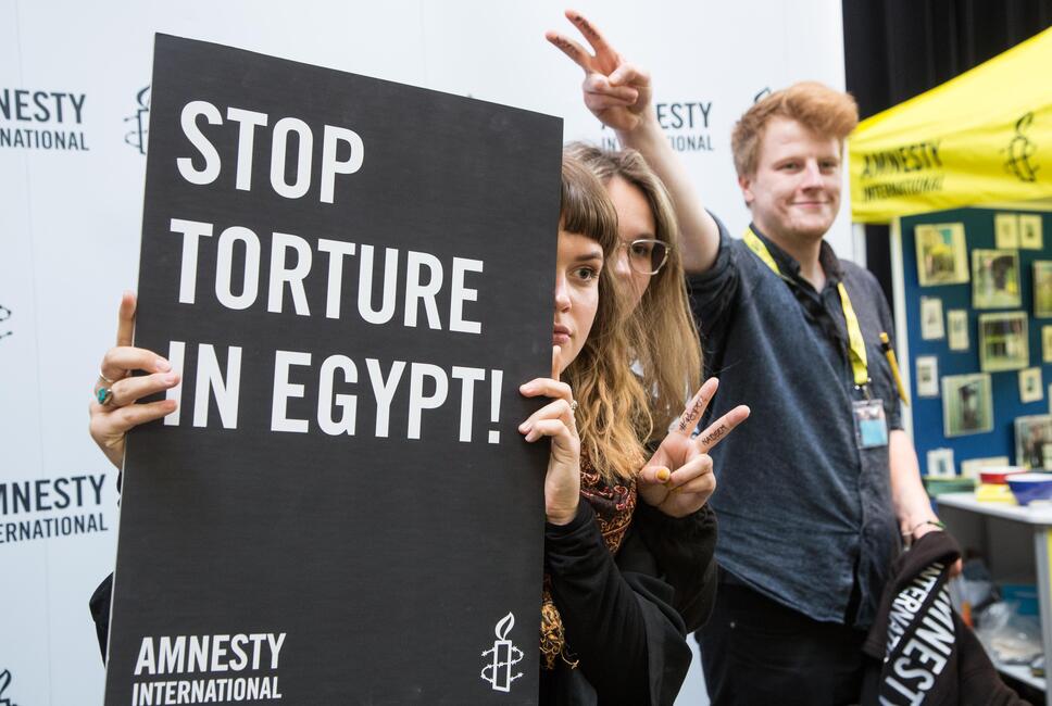 Zwei junge Frauen machen das Victory-Zeichen und halten ein Schild in die Kamera mit der Aufschrift:"Stop Torture in Egypt" Daneben steht ein junger Mann und macht auch ein Victory-Zeichen.