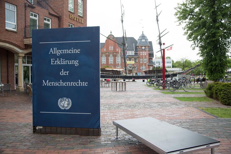 Das blaue Cover der "Allgemeine Erklärung der Menschenrechte" als Installation, aufgestellt auf einem Platz umgeben von Gebäuden aus rotem Backstein.