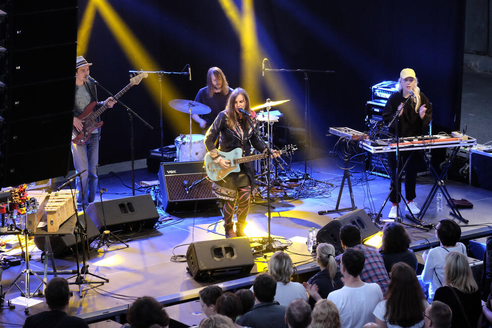 Eine Sängerin mit langem Haar und hellblauer Elektrogitarre spielt auf einer Bühne. Hinter ihr gelbes Scheinwerferlicht.