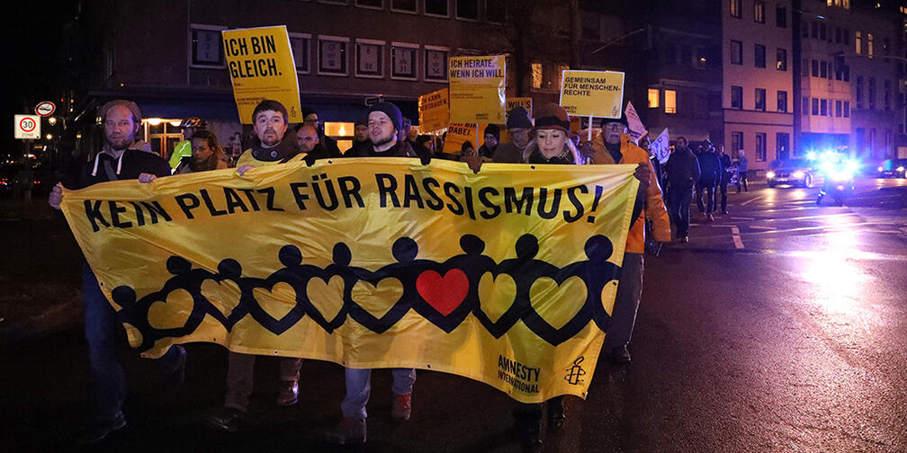 Viele Menschen tragen abends auf der Staße ein gelbes Banner vor sich mit dem Slogan "Kein Platz für Rassismus!"