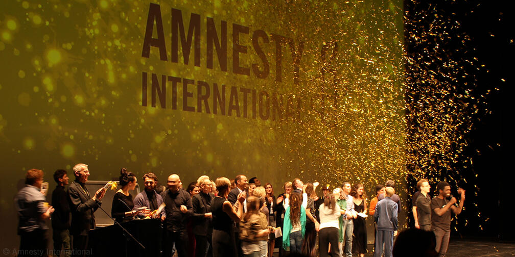 Eine Menschengruppe steht vor einer Leinwand, auf der groß Amnesty International steht