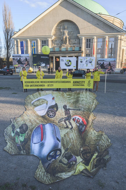 Menschen in gelben Amnesty-Jacken mit einem gelben Amnesty-Banner stehen vor einem Gebäude auf der Straße. Vor ihnen liegt ein großflächiges gemaltes Plakat, so dass der Eindruck entsteht, die Straße wäre aufgebrochen und unter ihr arbeiten Kinder an der Herstellung von Autos. 
