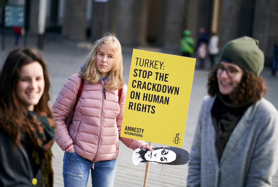 Drei junge Frauen stehen lächelnd im Kreis. Eine hält ein Pappschild mit der Aufschrift „Turkey: Stop the crackdown on human rights“