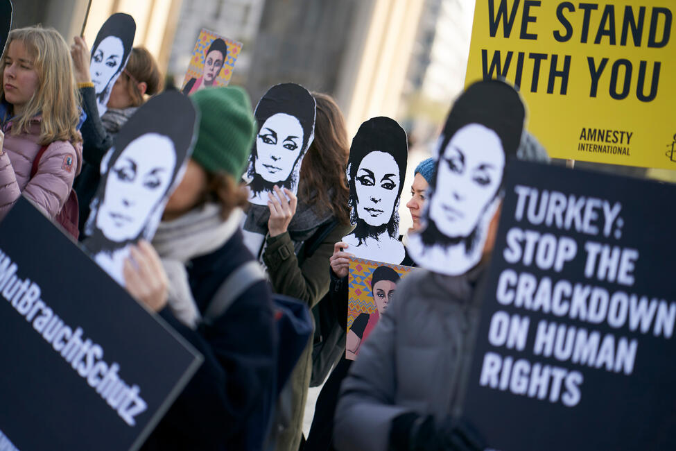 Eine Gruppe von Personen steht vor dem Brandenburger Tor und trägt Pappmasken mit dem Gesicht von Eren Keskin und hält Schilderin den Händen.