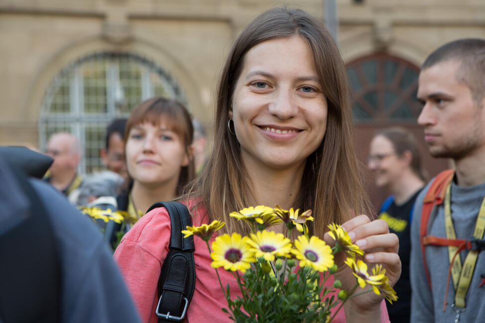 Eine junge Frau mit langen Haaren und gelben Blumen in der Hand lächelt in die Kamera. Hinter ihr andere Menschen.