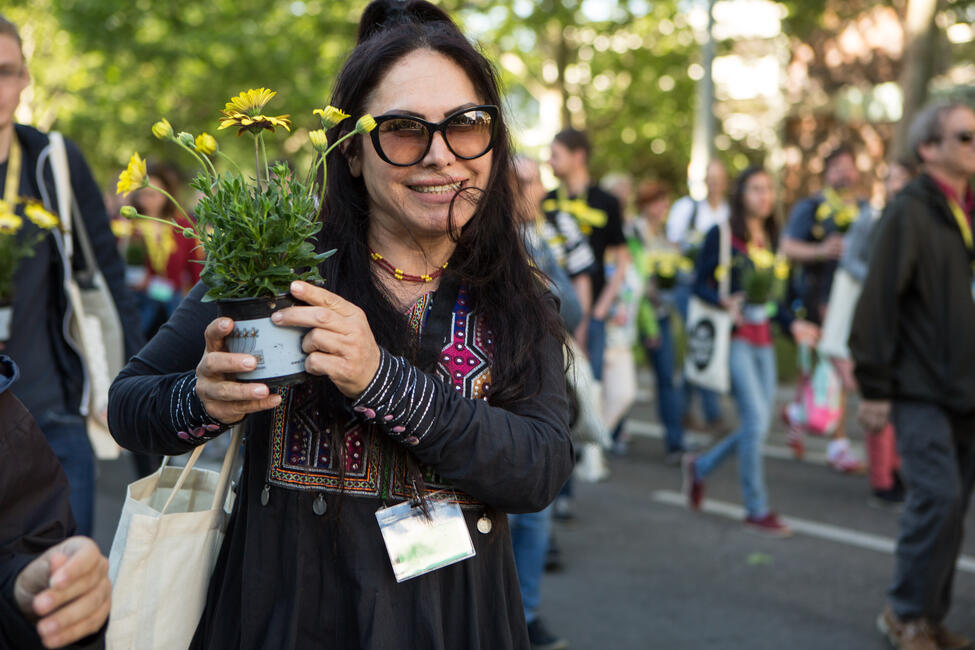 Eine junge Frau mit Sonnenbrille hält gelbe Blumen in einem Blumentopf in der Hand und ist Teil eine Demo