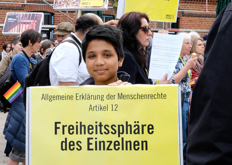 Junge guckt in Richtung der Kamera: Er hält ein gelbes Schild mit dem Text: "Freiheitssphäre des Einzelnen"