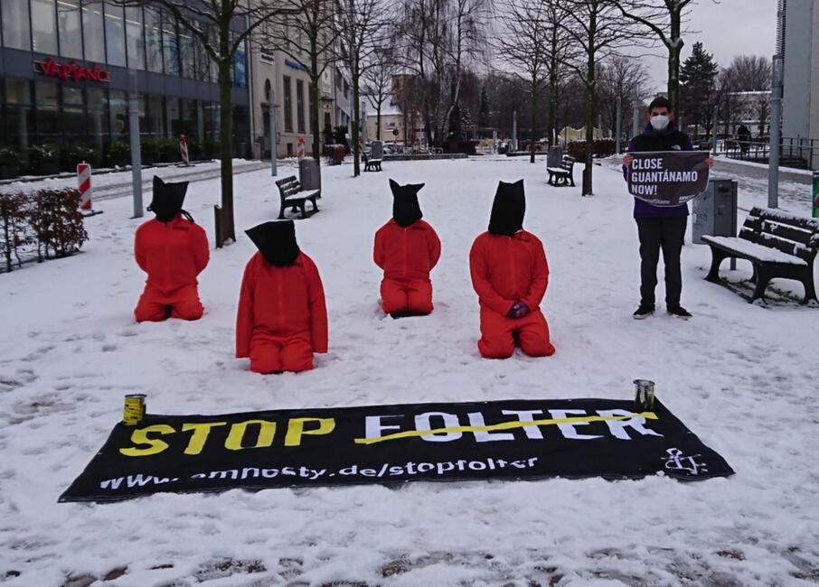 Das Bild zeigt vier Personen in orangenen Overalls mit schwarzen Säcken über den Köpfen, die im Schnee hinter einem Banner mit der Auufschrift "Stop Folter" knien. Neben ihnen steht ein Demonstrant mit Mundschutz, der ein Plakat mit der Forderung "Close Guantanamo now" hält.