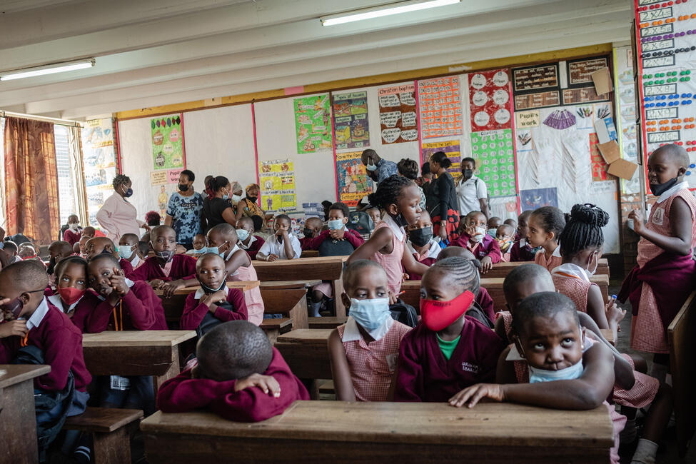 Afrikanische Schüler_innen sitzen an ihren hölzernen Schulbänken in einem Klassenzimmer, manche von ihnen tragen Mundnasenschutz.