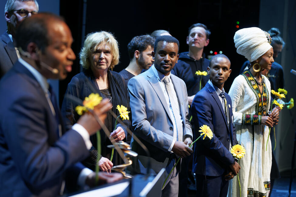 Das Bild zeigt mehrere Menschen auf einer Bühne, sie halten Blumen in der Hand