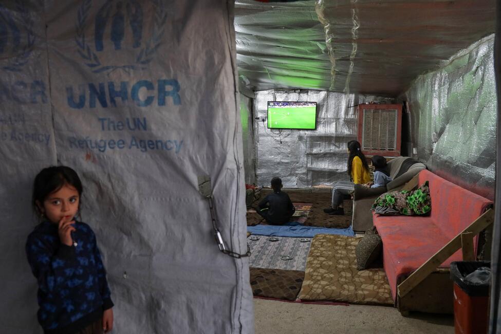 Ein Mädchen steht vor der Plane eines Zeltes in einem Flüchtlingslager, auf der Plane ist das Logo von "UNHCR" gedruckt, im Inneren des Zeltes gucken andere Kinder ein Fußballspiel auf einem Fernseher.