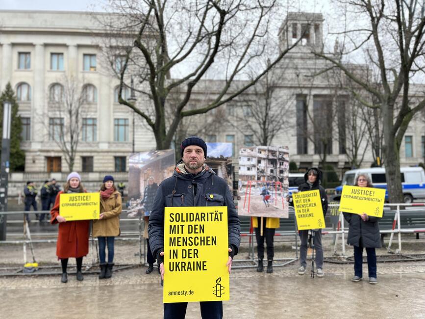 Das Bild zeigt mehrere Personen mit gelben Schildern, die vor einem Gebäude stehen.