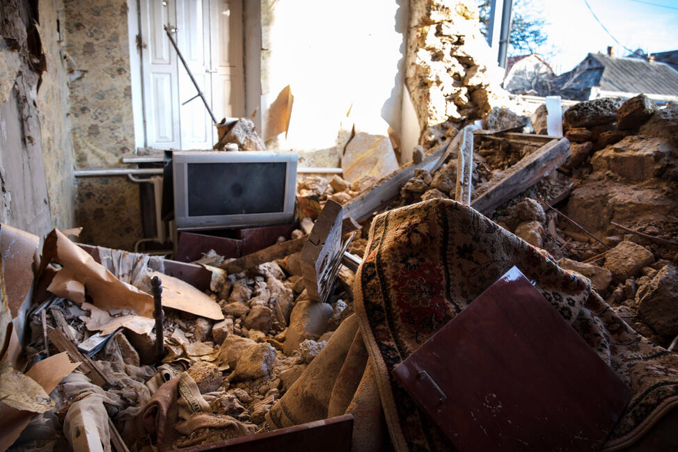 Das Foto zeigt ein total zerstörtes Wohnzimmer. Die rechts Wand fehlt. Steine und Schutt liegen über den Möbeln. Ein Fernseher ist mit Staub bedeckt.