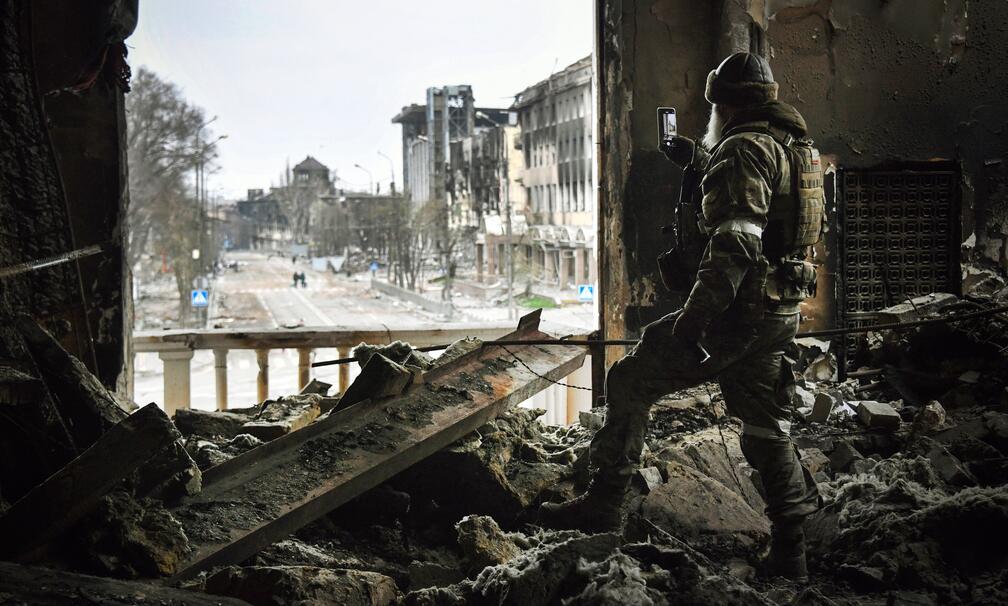 Das Bild zeigt eine bewaffnete Person in Armee-Kleidung, die in einem stark zerstörten Gebäuade steht und mit dem Handy nach draußen auf eine verwüstete Straße fotografiert.