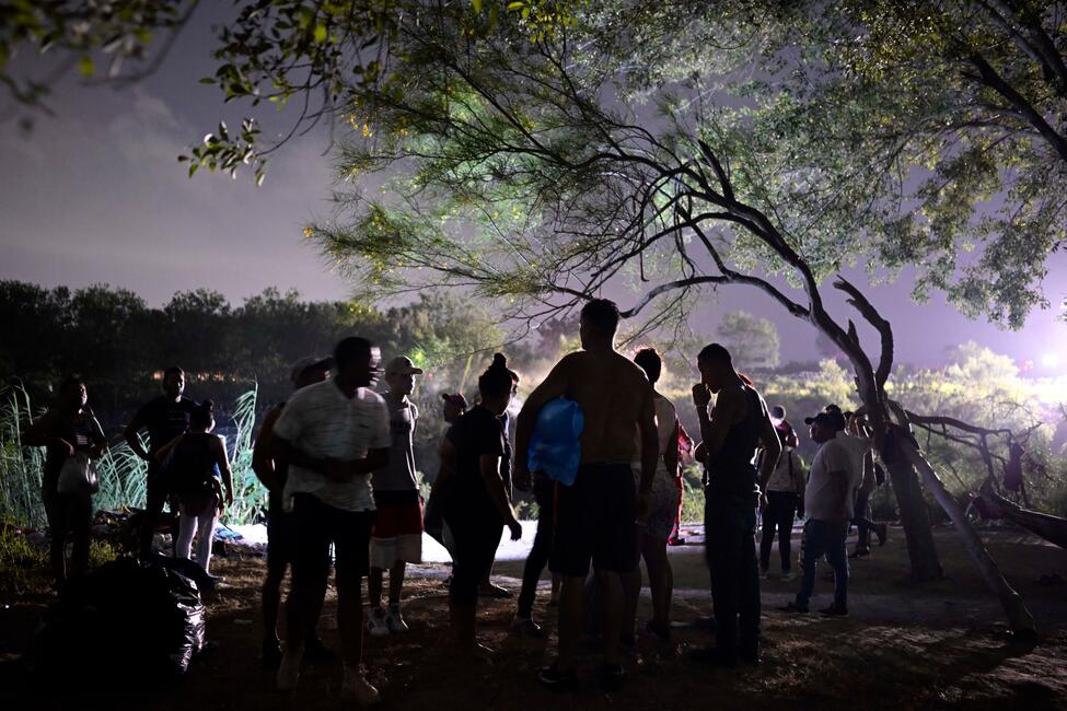 Menschen versammeln sich in der Natur bei Nacht unter einem Baum, ein Scheinwerfer strahlt aus der Ferne, sie tragen T-Shirts und kurze Hosen oder lange Jeans, manche Rucksäcke.