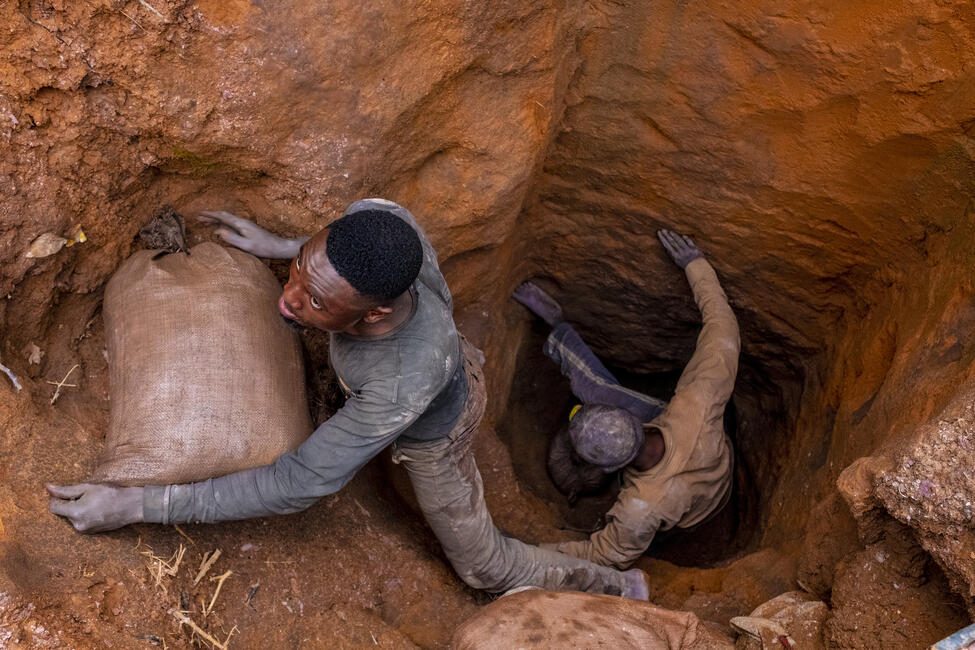 Zwei Männer entsteigen einem Erdloch, das in eine Mine führt, einer von ihnen hat einen prallgefüllten Stoffsackin den Armen.