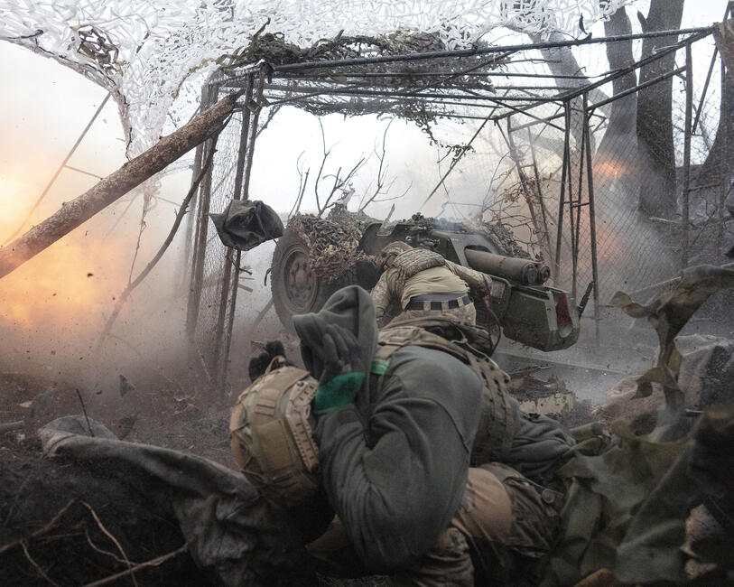Soldat*innen im Gefecht, sie schießen etwas ab, eine Explosion, ein Soldat geht in Deckung unter seinem Helm, hält sich die Ohren zu, über ihm und anderen Soldat*innen aufgespannt: Tarnnetze.