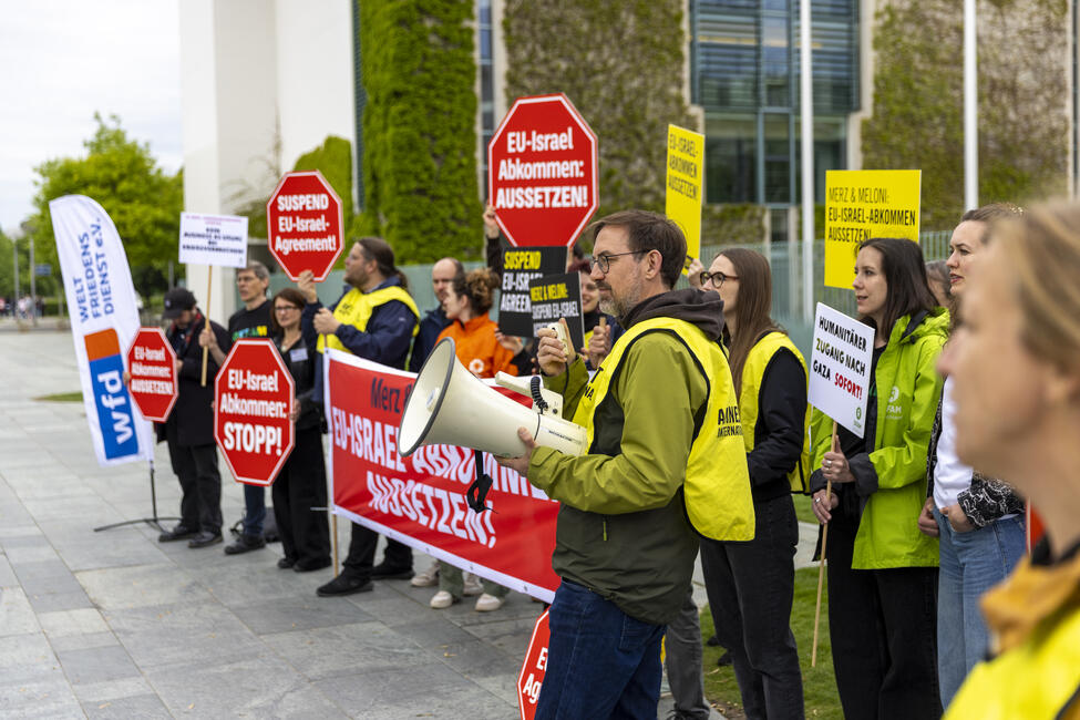 Menschen bei Protestaktion vor dem Kanzleramt in Berlin mit Transparenten und Schildern. Eine Person hält ein Megaphon.