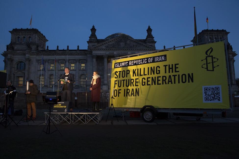 Das Bild zeigt den Deutschen Bundestag bei Nacht, davor steht ein Schild auf einem Wagen mit dem Schriftzug "Stop killing the future generation of Iran". Daneben stehen mehrere Personen, von denen eine in ein Mikro spricht.