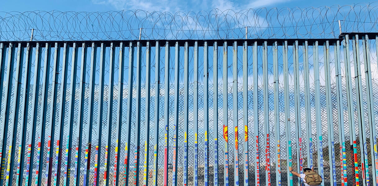 Ein meterhoher Grenzzaun aus Stahlstreben, darüber Stacheldrahtzaun, eine Person stemmt sich gegen den Zaun