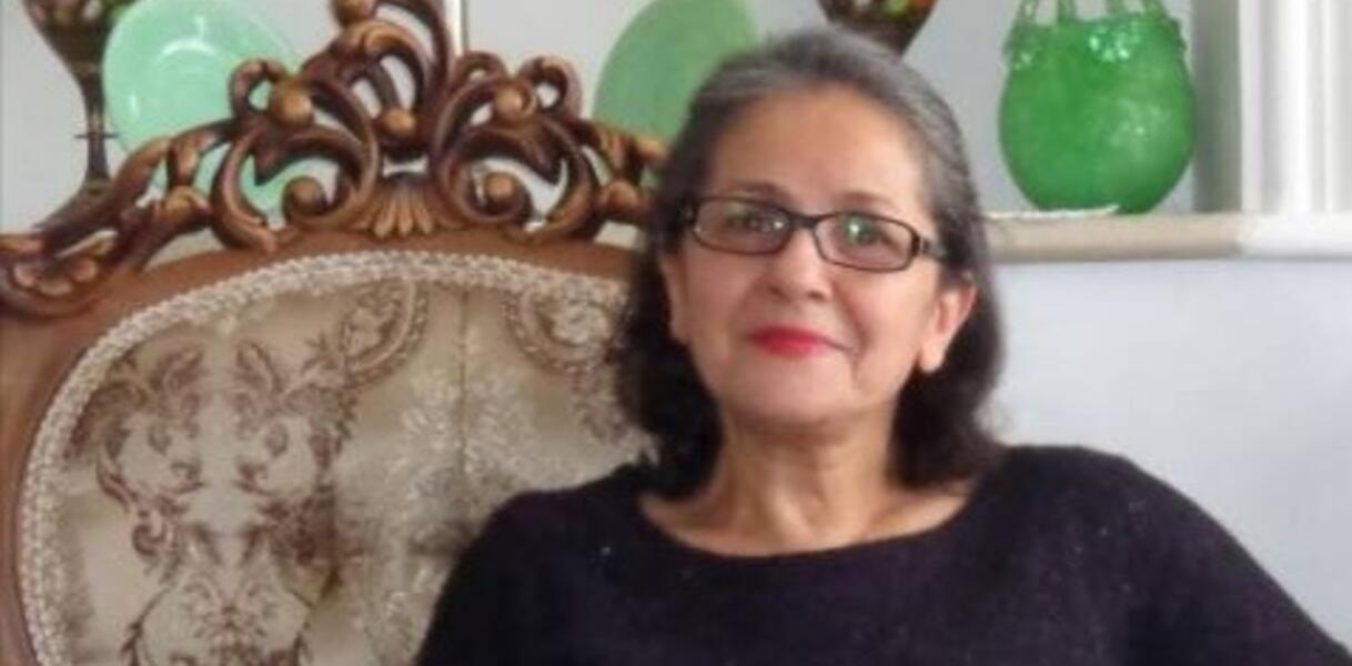 Eine ältere Frau mit schulterlangen grauen Haaren sitzt auf einem eleganten Sessel. Sie trägt eine Brille, roten Lippenstift und ein schwarzes Kleid.