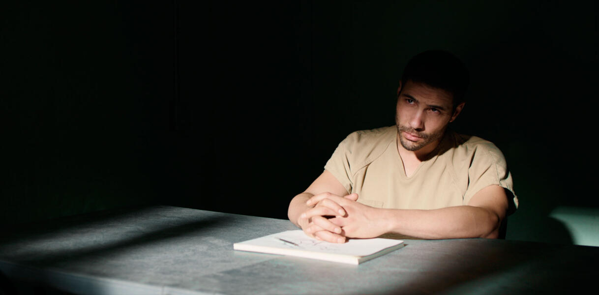 Ein Mann mit kurzem Bart in einem Häftlingsanzug sitzt im Halbdunkeln an einem Tisch.