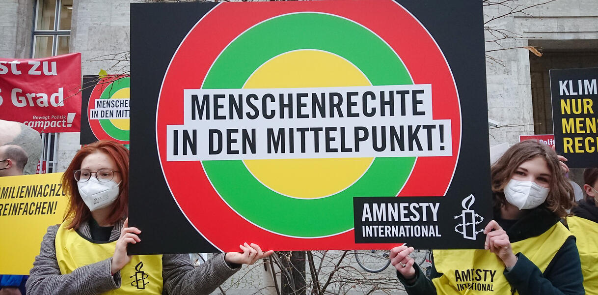 Das Bild zeigt mehrere Menschen mit Protestplakaten, auf einem steht "Menschenrechte in den Mittelpunkt"