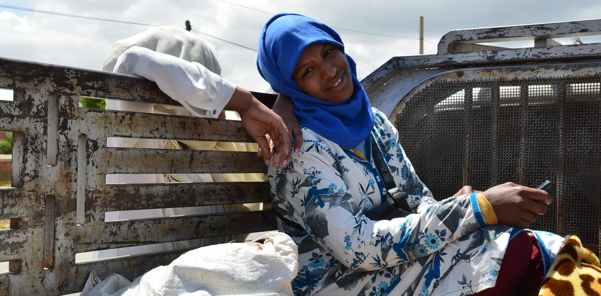 Ein junges Mädchen mit einem Kopftuch sitzt in marokkanischer Kleidung auf der Ladefläche eines Lasters und lächelt, während sich eine Person mit ihr unterhält.
