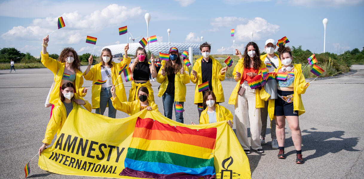 Menschen in gelben Jacken wedeln mit kleinen Regenbogenflaggen. Im Hintergrund ist die Allianz-Arena zu sehen, im Vordergrund halten mache der Menschen ein gelbes Banner mit einer großen Regenflagge drauf und mit der Aufschrift "Amnesty International". 