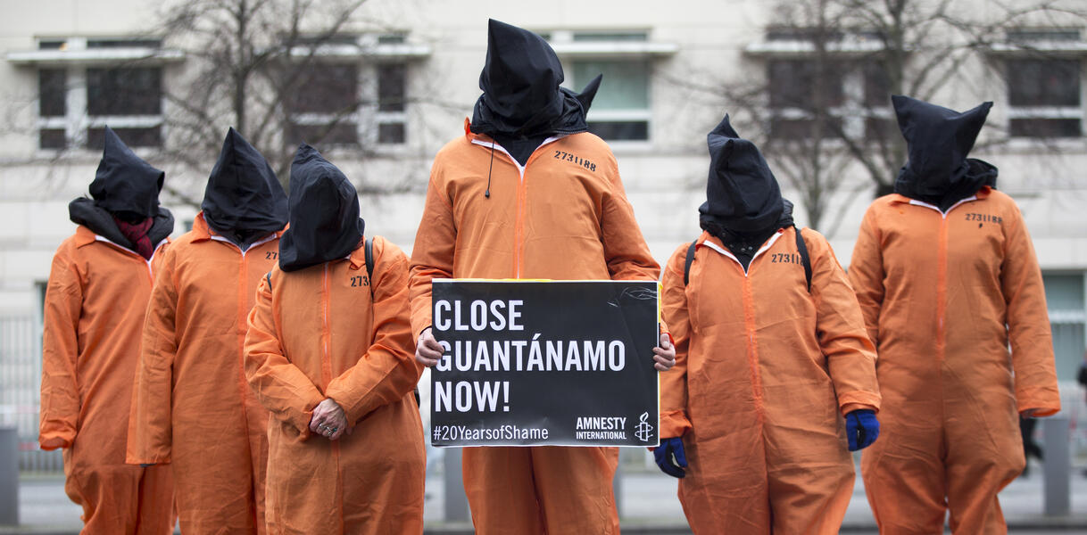Das Bild zeigt sechs Menschen in orangenen Overalls mit schwarzen Säcken über den Köpfen. Eine Person hält ein Schild mit der Aufschrift "Close Guantanamo now!".