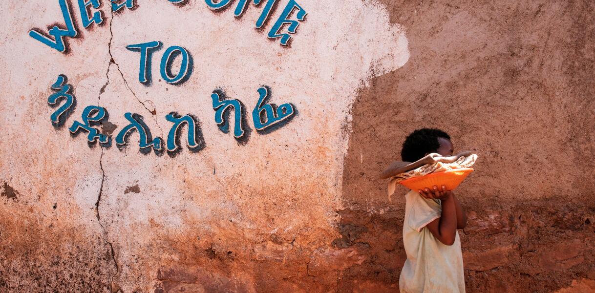 Das Bild zeigt ein Schwarzes Kind, das eine Schüssel mit Tüchern trägt und an einer Wand mit einem Willkommens-Schriftzug entlang läuft.