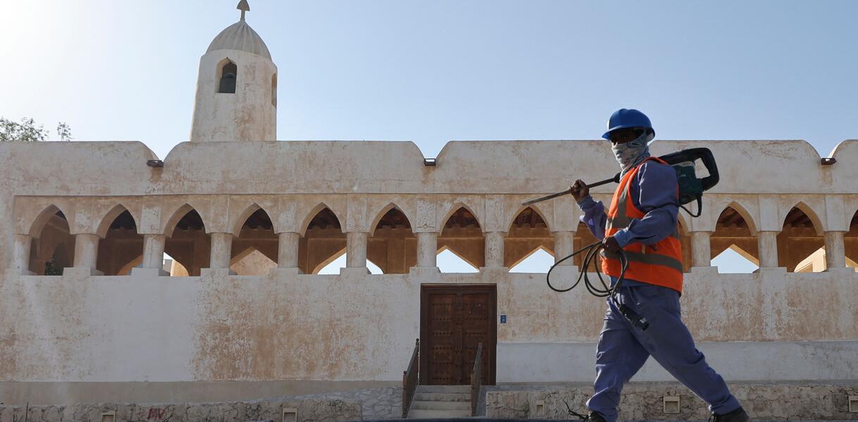 Ein Arbeiter in Arbeitskleidung und mit einem Werkzeug vor einem traditionellem Gebäude.
