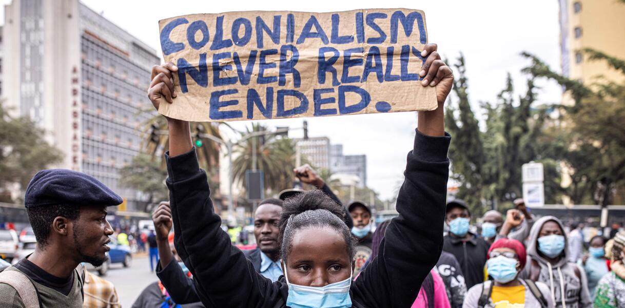 Eine Frau mit Maske hält ein Schild in die Höhe auf dem steht "Kolonialism never really ended". Im Hintergrund laufen weitere Menschen.