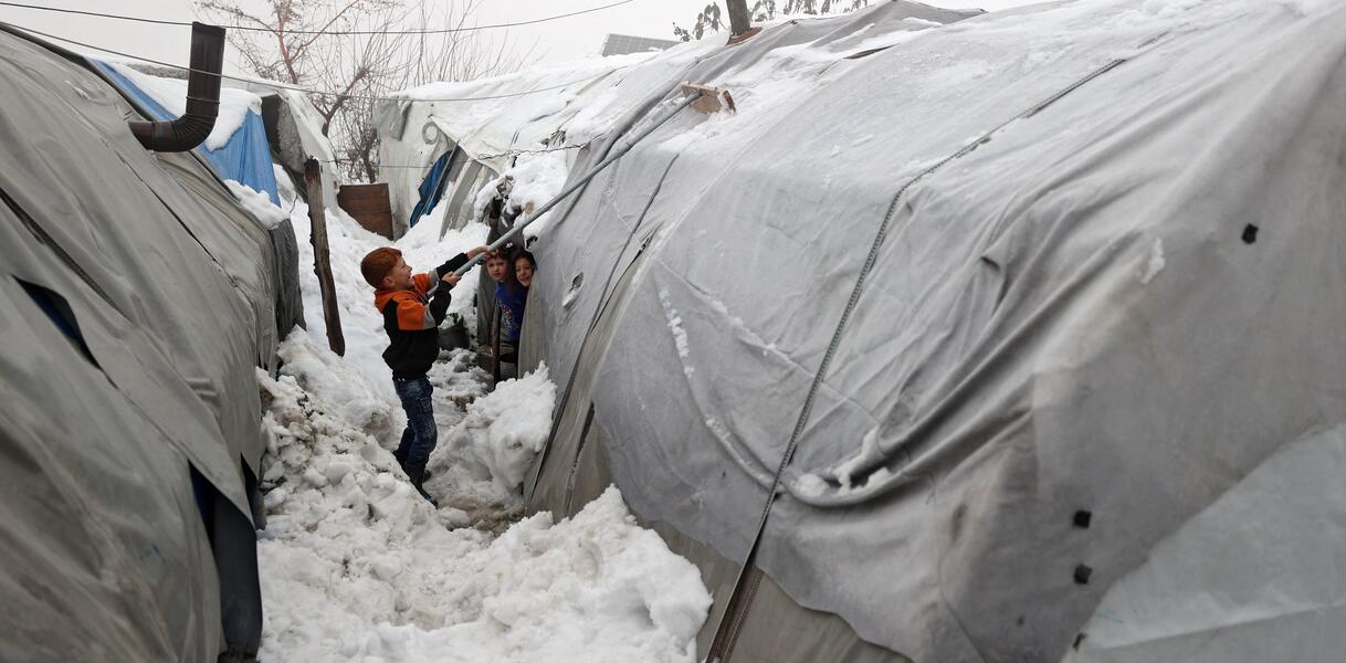 Ein Junge steht inmitten großer Schneemassen zwischen zwei Zelten und entfernt mit einem Besen Schnee von einem der Zelte. Zwei weitere Kinder stehen neben einem der Zelte und schauen ihm dabei zu.