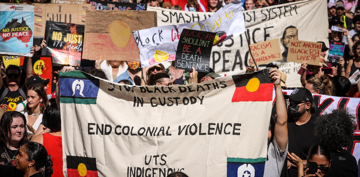 Demonstration mit Schildern und Bannern. Zentral zeigt das Bild ein Banner mit der Aufschrift "End colonial violence", also "Koloniale Gewalt beenden".