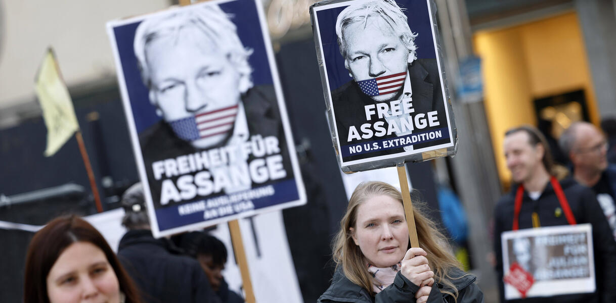 Das Bild zeigt zwei Personen, die demonstrieren. Sie haben Schilder mit dem Foto von Julian Assange in der Hand