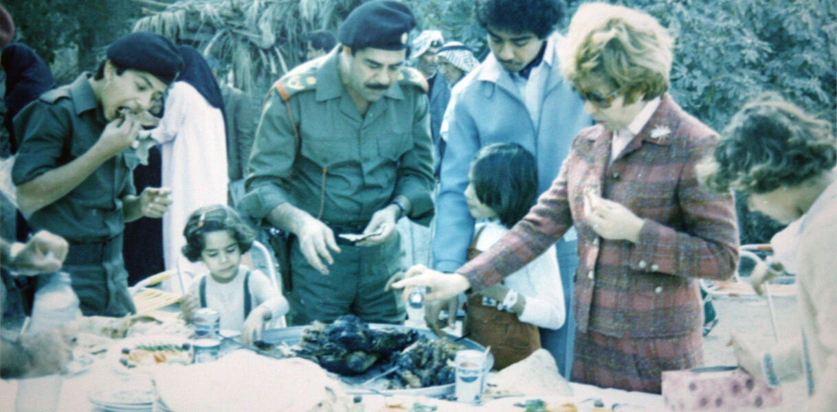 Der Diktator Saddam Hussein steht zusammen mit Mitgliedern seiner Familie an einem Buffet, das draußen aufgebaut ist, und bedient sich.