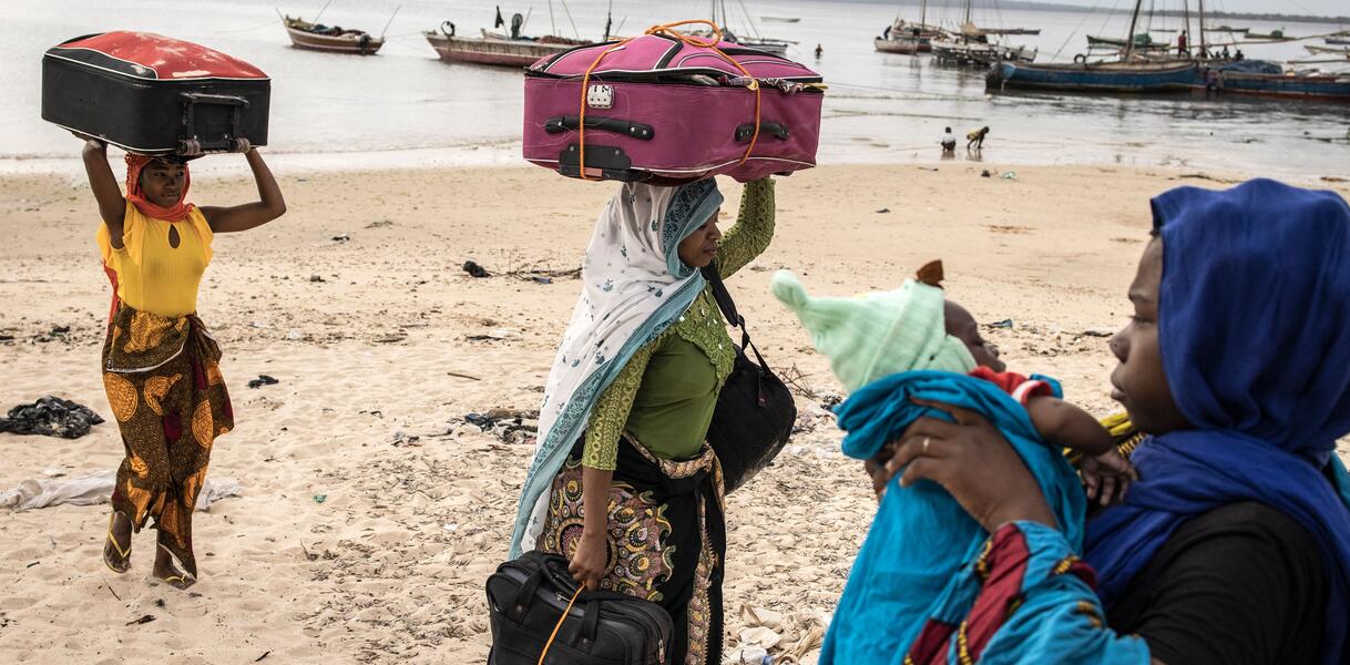 Das Bild zeigt drei Frauen und ein Baby mit Koffern an einem Strand.