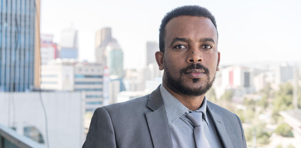 Ein äthiopischer Mann mit kurzen Haaren und kurzem Bart trägt ein Hemd und Jacket und Krwatte, steht vor der Skyline einer Großstadt, ernste Mimik. 