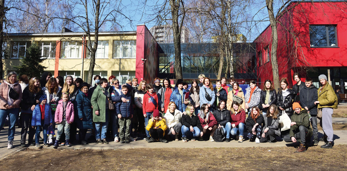 Kinder einer Schulklasse haben sich zusammen mit ihren Lehrerinnen zum Foto aufgestellt, hinter ihnen stehen Bäume und Schulgebäude.Schüler_innen und Lehrerinnen aus Charkiw vor der Migrationsbehörde in Vilnius aufgestellt.