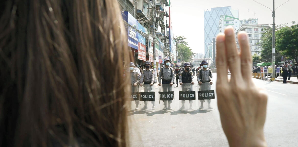 Eine demonstrierende Person mit langen Haaren hält drei Finger der rechten Hand hoch und steht dabei einer Gruppe von Sicherheitskräften gegenüber, die Helme tragen und  Schilder, auf denen "Police" geschrieben steht.