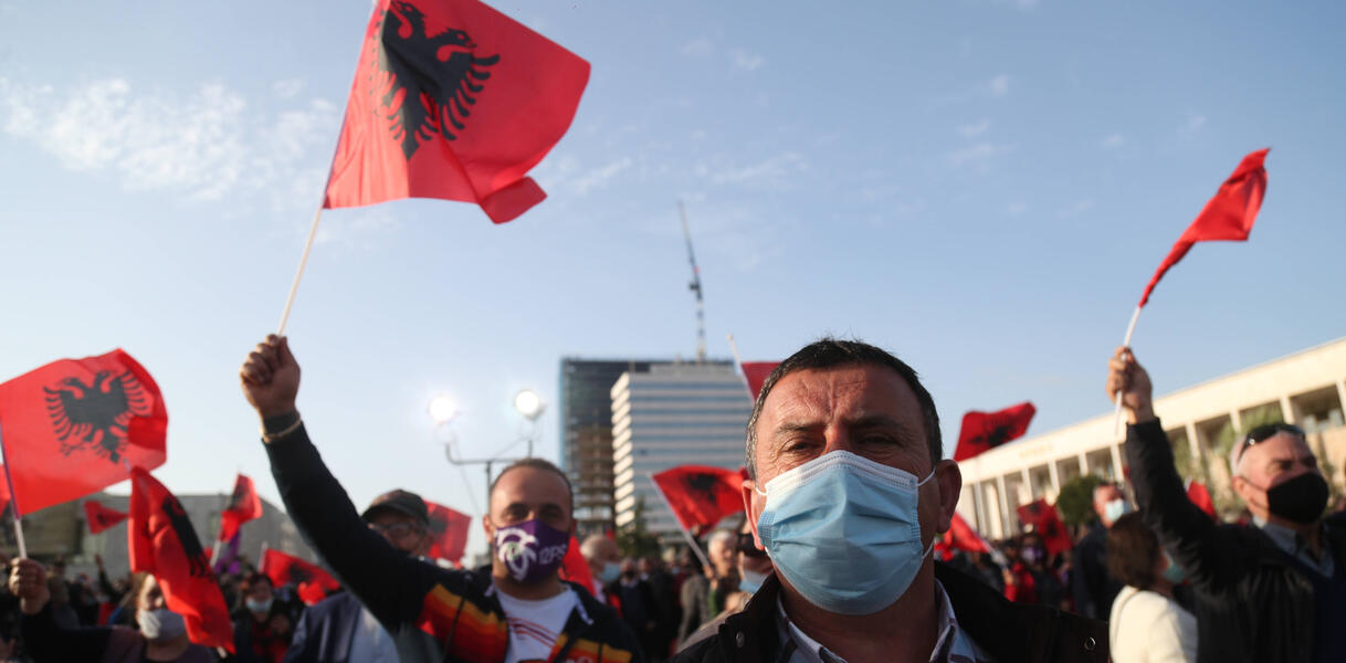 Viele Meschen mit Masken schwenken albanische Flaggen vor blauem Himmel.